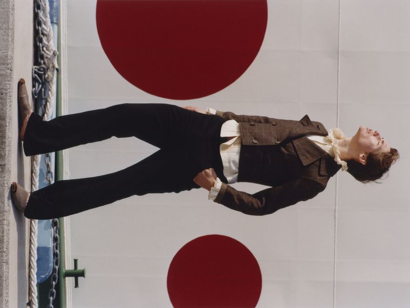 Model wearing a brown tweed jacket and dark trousers standing against a wall with large red circular patterns for ODDA Magazine