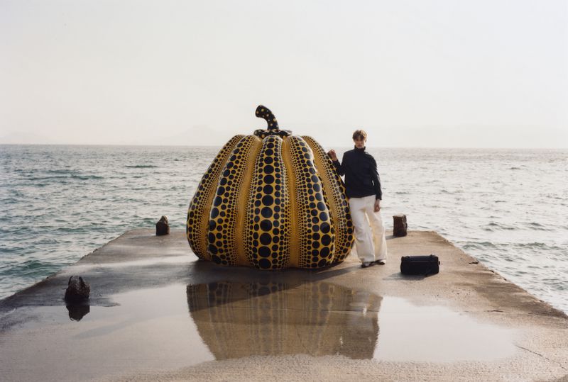 Man standing on a concrete pier leaning against a large yellow and black polka dot pumpkin sculpture by the ocean.