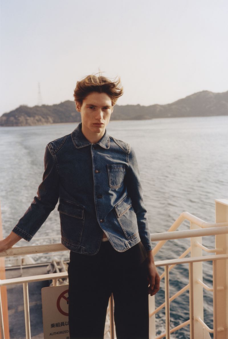 Male model in a denim jacket leaning on a ferry railing with a coastal landscape background