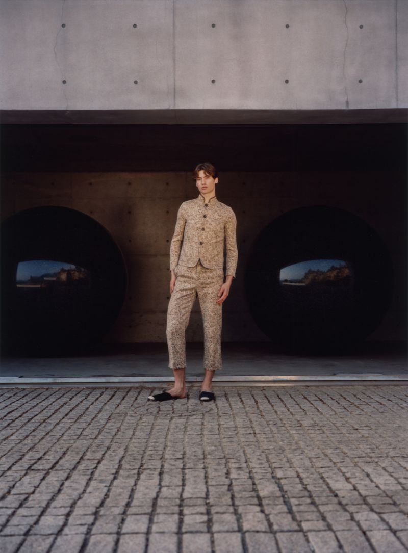 A young male model standing on a cobblestone courtyard wearing patterned matching jacket and trousers against a concrete architectural backdrop.