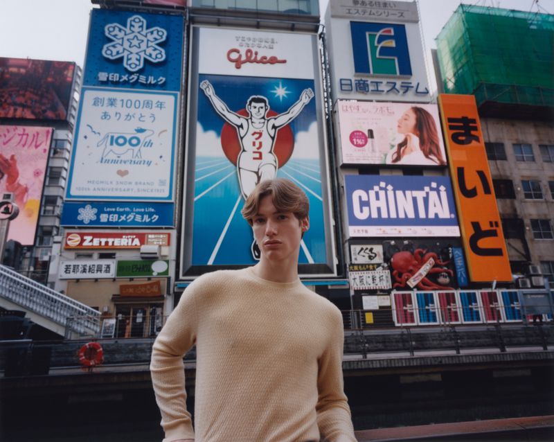 Young man wearing a cream sweater posing in front of brightly lit neon billboard signs in a city street.