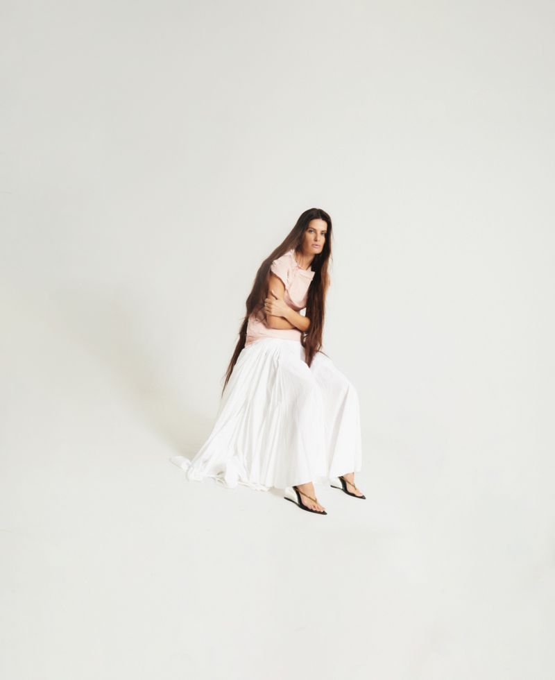 Model Isabeli Fontana seated against a white background wearing a pink top and long white pleated skirt.