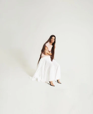 Model Isabeli Fontana seated against a white background wearing a pink top and long white pleated skirt.