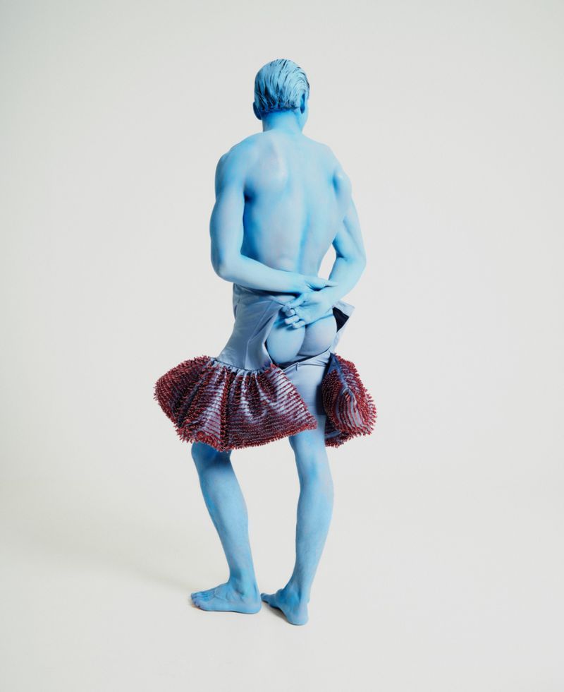 Full-length rear view of a blue-painted man wearing a ruffled skirt against a seamless white studio background.