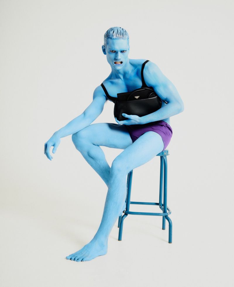 Man with blue body paint poses with black bag, sitting on a stool in a studio setting, wearing purple shorts and a black top.