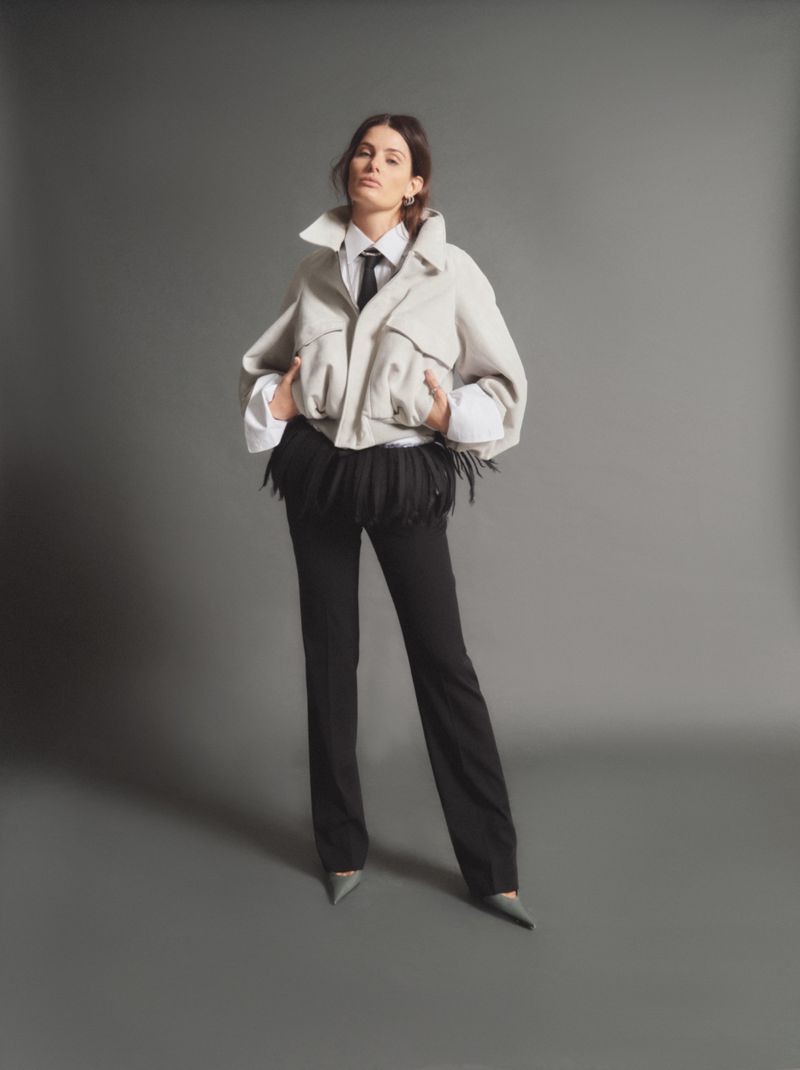 Model Isabeli Fontana wearing a white jacket over a white shirt and black pants against a solid grey studio background.