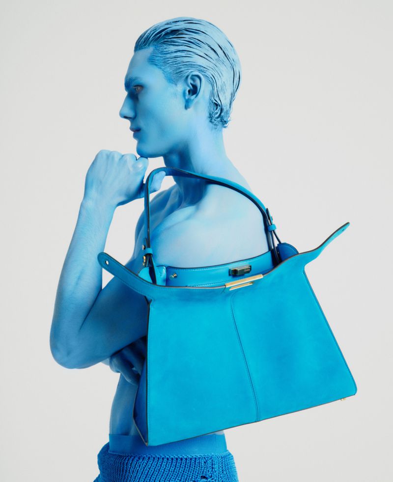 Monochrome blue painted model holding a bright blue leather handbag against a pale grey seamless background