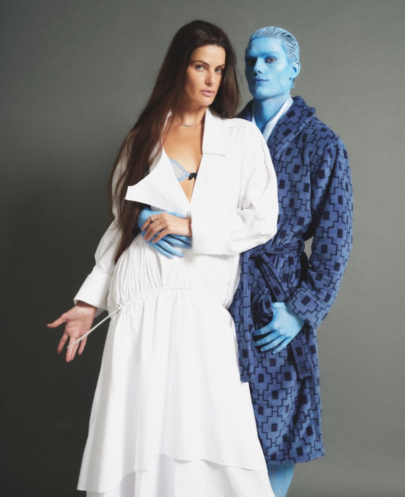 Model Isabeli Fontana in a white dress standing beside a blue-painted male model in a patterned robe against a grey backdrop.