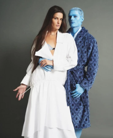 Model Isabeli Fontana in a white dress standing beside a blue-painted male model in a patterned robe against a grey backdrop.