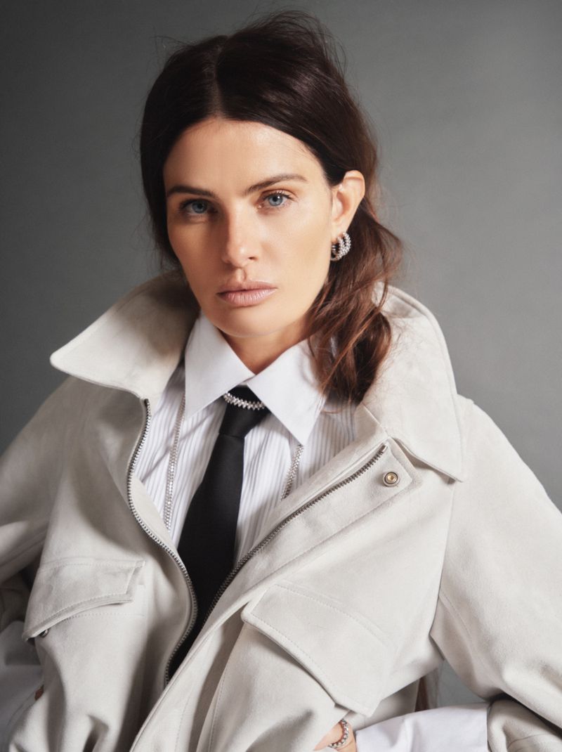 Model Isabeli Fontana wearing a light jacket, white collared shirt, and black tie against a grey background