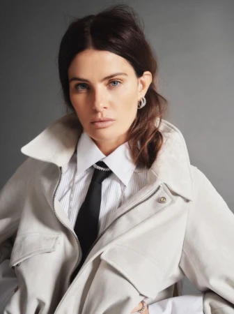 Model Isabeli Fontana wearing a light jacket, white collared shirt, and black tie against a grey background