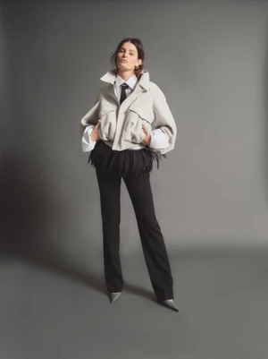 Model Isabeli Fontana wearing a white jacket over a white shirt and black pants against a solid grey studio background.
