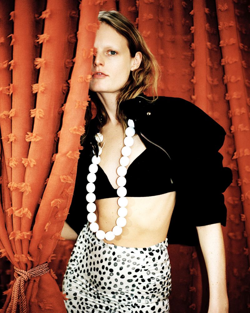 Hanne Gaby Odiele partially hidden by an orange curtain wearing a black jacket and white beaded necklace