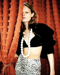 Hanne Gaby Odiele partially hidden by an orange curtain wearing a black jacket and white beaded necklace