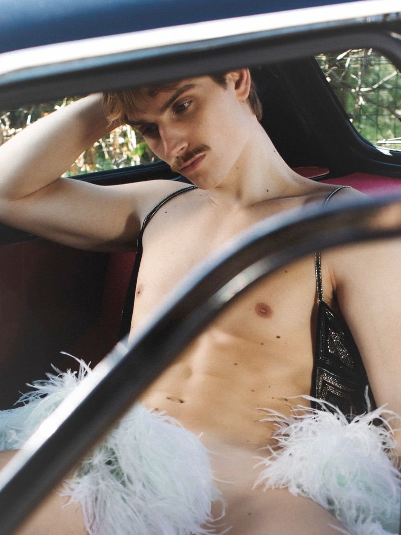 A young male model with a mustache seated inside a vintage car wearing a feathered garment