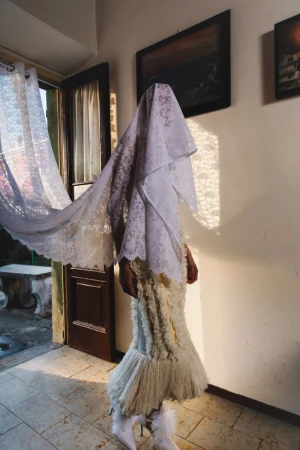 Model draped in a lace veil wearing a textured white gown inside a domestic interior near an open doorway.