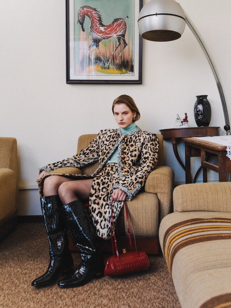 Model in leopard print coat and tall black boots seated on an armchair beneath a framed horse painting