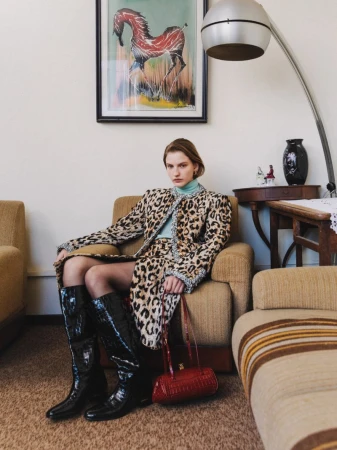 Model in leopard print coat and tall black boots seated on an armchair beneath a framed horse painting