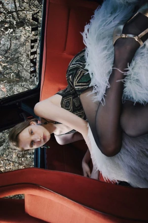 Model reclining in the back seat of a classic car with bright red interior, wearing a patterned dress and feathery skirt