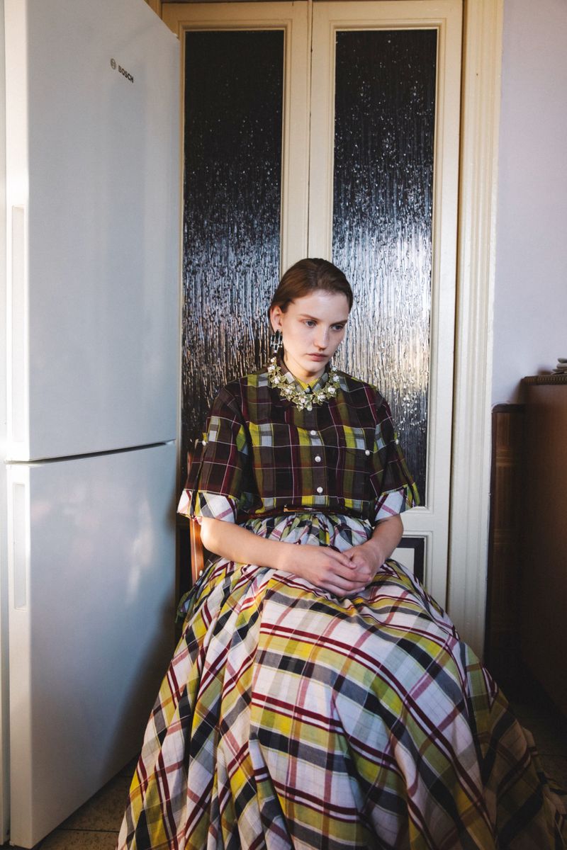 Seated woman in a plaid dress and ornate collar indoors next to a white refrigerator.