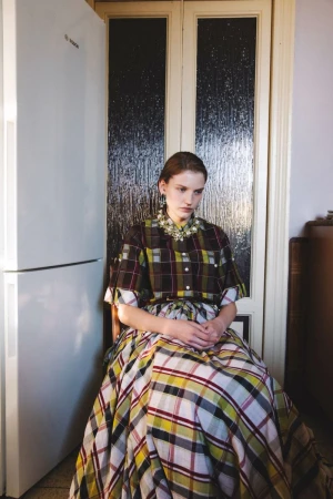 Seated woman in a plaid dress and ornate collar indoors next to a white refrigerator.