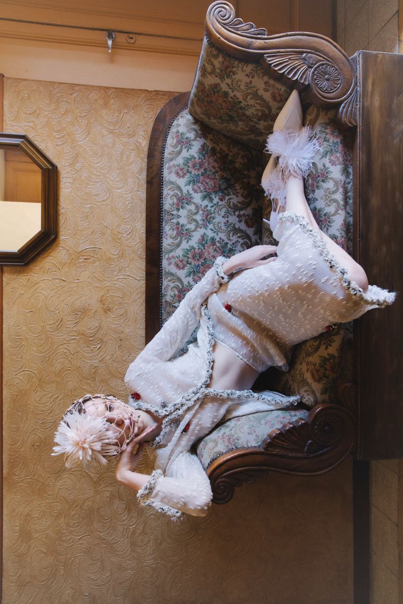 Model lying on an antique patterned sofa wearing an intricate textured white outfit and feathered footwear.