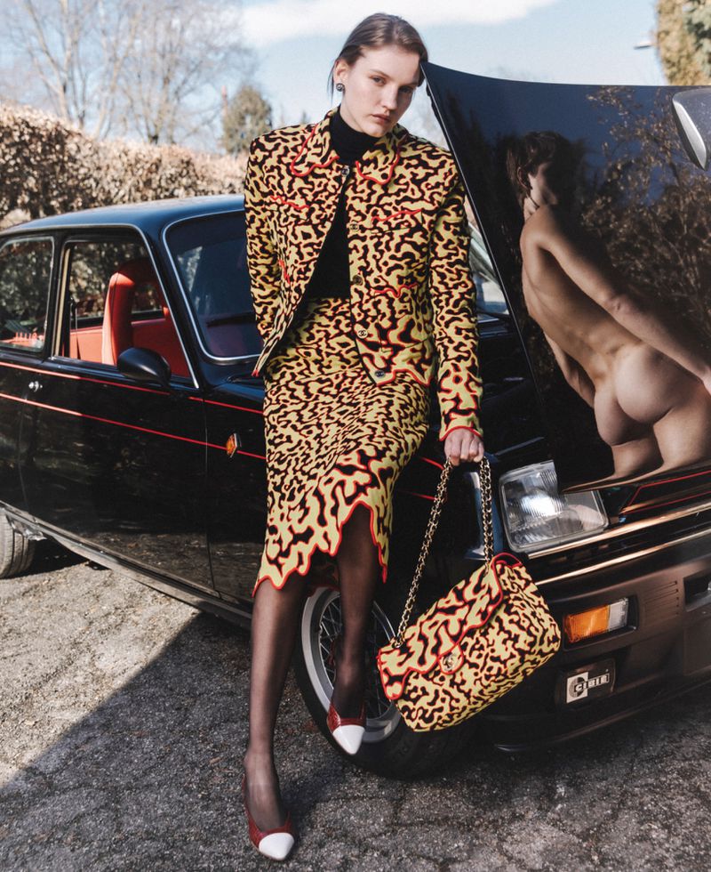 Model leaning against a black vintage car wearing a yellow patterned jacket, skirt, and holding matching handbag