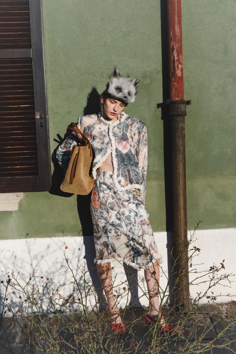 Model wearing patterned jacket and skirt, animal hat, and holding tan handbag against a green wall