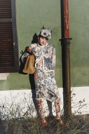 Model wearing patterned jacket and skirt, animal hat, and holding tan handbag against a green wall