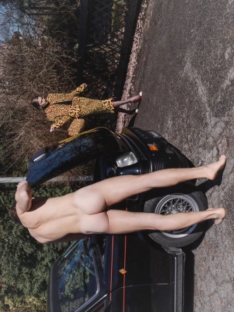 A nude person leaning over a black car with a second person wearing a patterned yellow outfit standing nearby on an asphalt surface.
