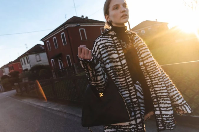 Model in a black and white patterned coat carrying a black shoulder bag outdoors at sunset with buildings in the background.