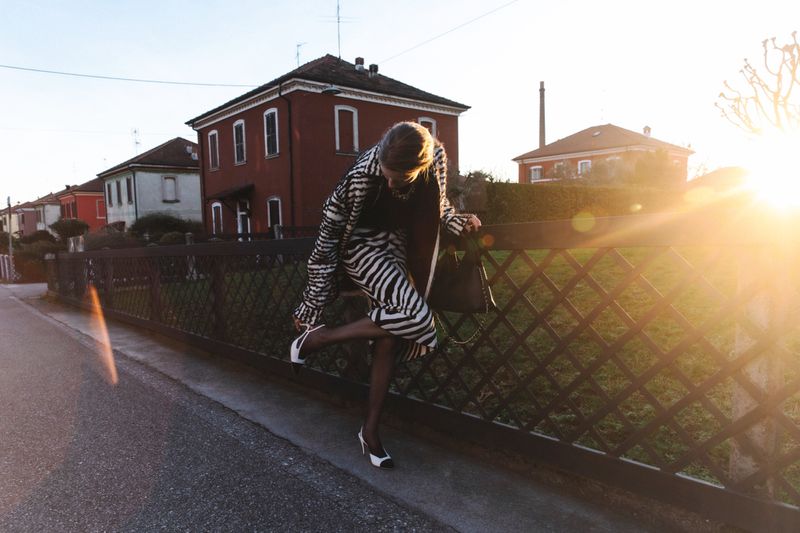 Model in zebra-print coat adjusting heels outdoors near a wooden fence at sunset for ODDA Magazine