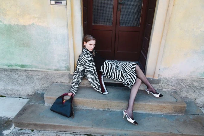 Model seated on stone steps in front of a wooden door wearing a zebra print coat and skirt holding a black handbag