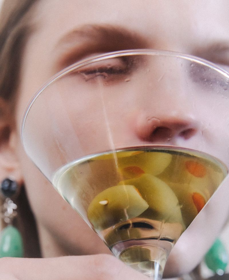 Close-up portrait of a woman holding a martini glass containing olives, with green gemstone earrings visible.