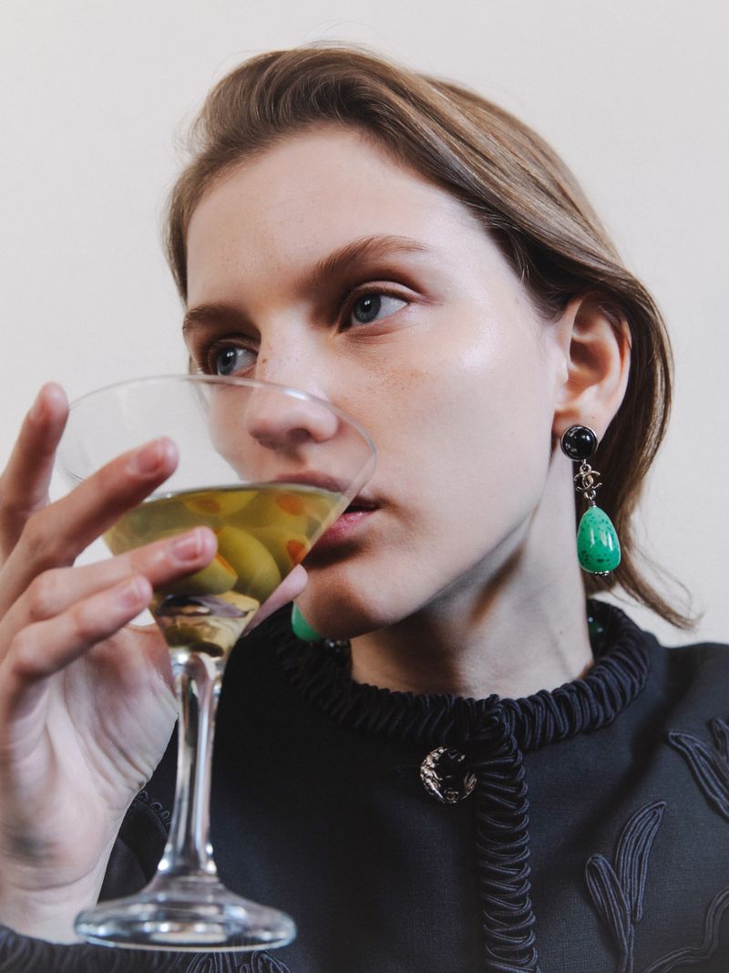 Close-up of a woman in a black top holding a martini glass with olives against a plain light background.