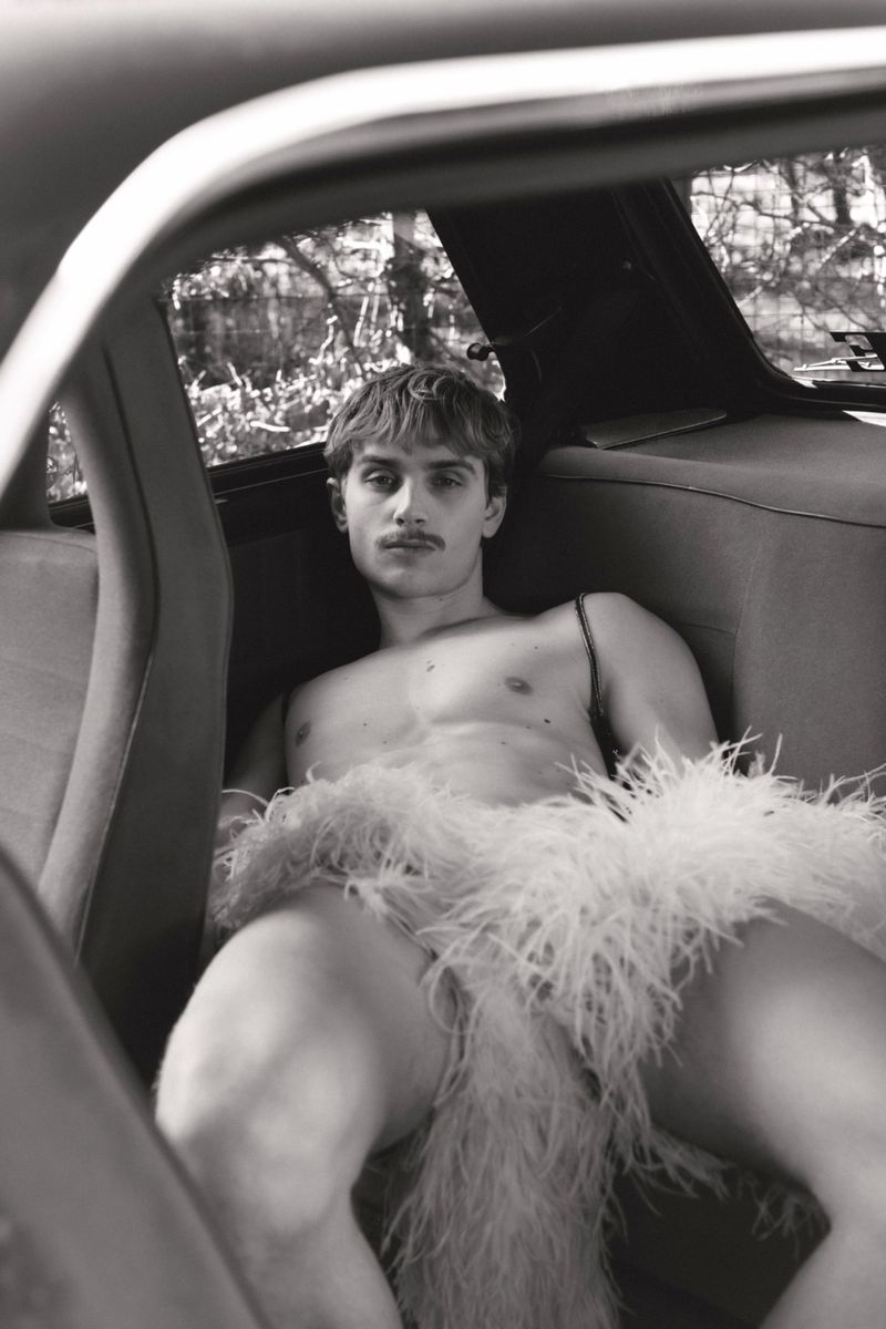 Black and white portrait of a man with a mustache seated inside a car wearing a feather garment.