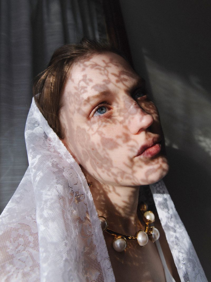 Close-up portrait of a woman wearing a white lace head covering and pearl necklace with dappled light on her face.