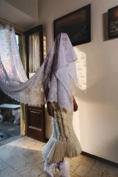 Model draped in a lace veil wearing a textured white gown inside a domestic interior near an open doorway.
