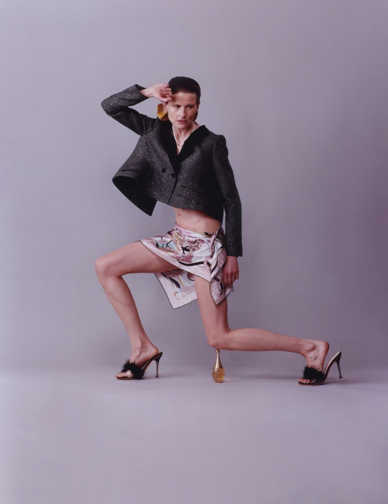 Model posing in a structured jacket and patterned wrap skirt against a plain grey studio background.