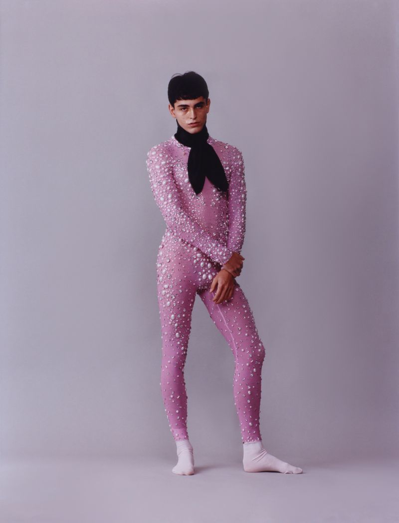 Model in a pink embellished bodysuit and black neck scarf standing against a plain grey studio background.