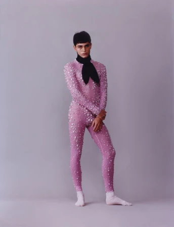 Model in a pink embellished bodysuit and black neck scarf standing against a plain grey studio background.