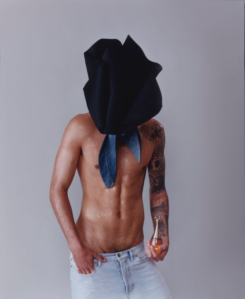 Shirtless tattooed man wearing a large black sculptural headpiece holding a perfume bottle against grey seamless background.