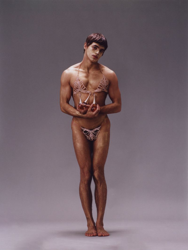 A shirtless male model standing against a grey studio backdrop, wearing delicate beaded underwear and holding two glass perfume bottles.