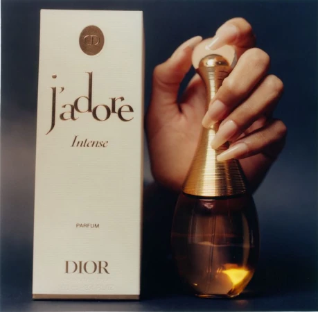 Hand with long manicured nails holding a perfume bottle next to its branded white box against a dark background.