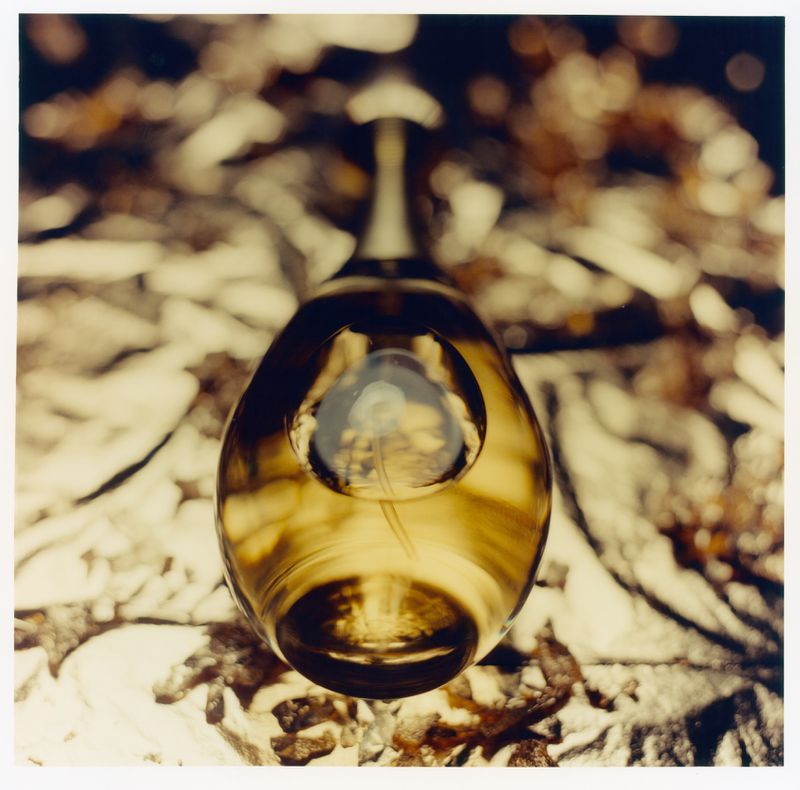 An amber glass perfume bottle rests on a textured metallic surface, captured from a high-angle macro perspective.