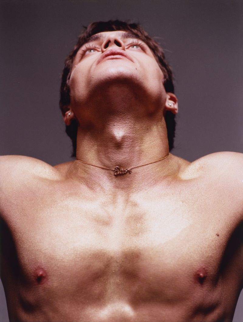 Close-up portrait of a male model looking upward with a delicate chain necklace against a dark grey studio background.