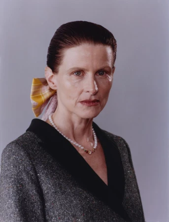Portrait of an older woman wearing a tweed jacket, pearl necklace, and a patterned hair ribbon against a plain grey background