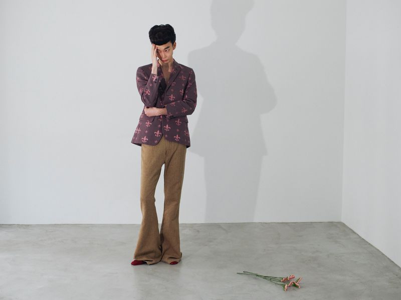 Man standing in patterned purple blazer and brown trousers touching his head against a white studio corner wall background.