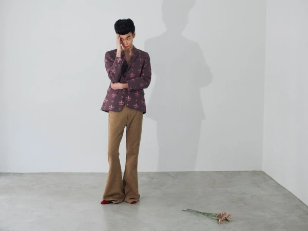 Man standing in patterned purple blazer and brown trousers touching his head against a white studio corner wall background.