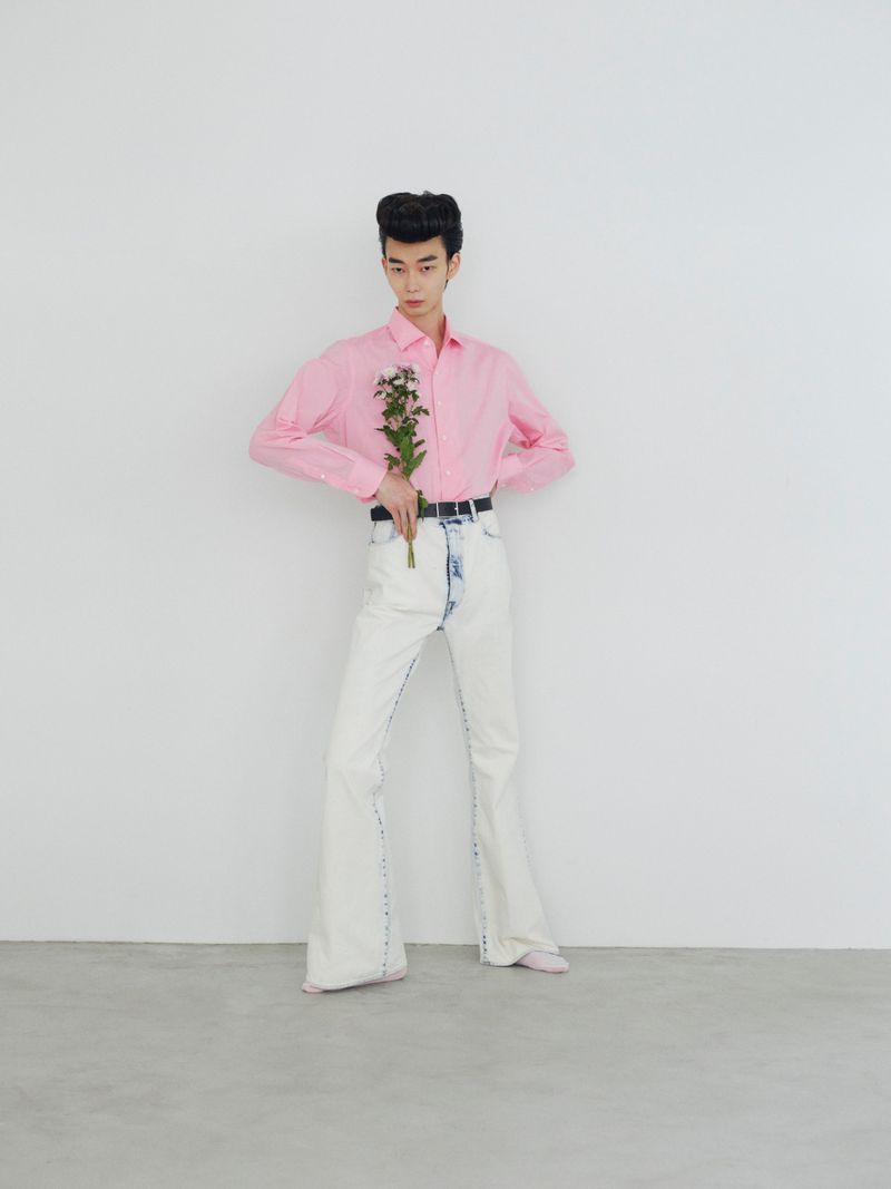 Model wearing a pink long-sleeve shirt and white pants holding a small bouquet of flowers against a seamless backdrop